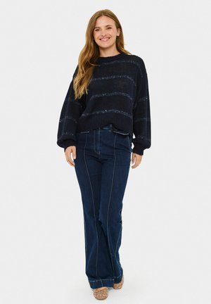 Granatowy sweter dzianinowy z poziomymi metalicznymi paskami, luźny krój, okrągły dekolt, zestawiony z ciemnymi dzwonami i jasnobrązowymi sandałami na obcasie.