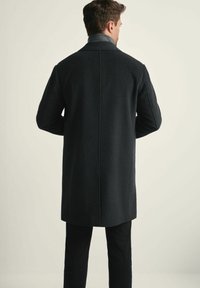 Mężczyzna widziany z tyłu, ubrany w ciemnoszary płaszcz i czarne spodnie, stojący na jednolitym jasnym tle.