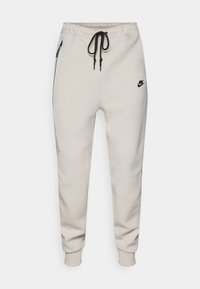 Ljusbeige Nike-joggers med elastisk midjeband, svart dragsko och sidofickor med blixtlås. Har en liten svart logga på framsidan.