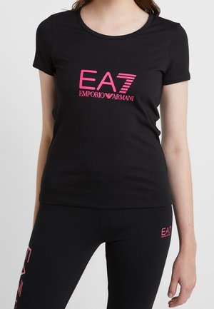 Kobieta ubrana w czarny top z krótkim rękawem i legginsy z różowym logo EA7 Emporio Armani na klatce piersiowej, udzie i łydce na jednolitym jasnym tle.