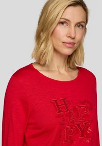 Rotes Langarmshirt mit Rundhalsausschnitt, das metallisch bedruckte Buchstaben mit dem Text "HAPPY MY STATE OF MIND" in unterschiedlichen Größen zeigt. Weiche Textur.