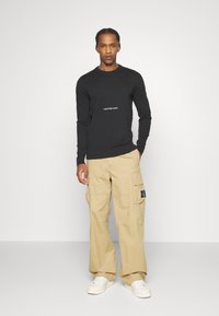 Maglione nero a maglia con scollo rotondo, caratterizzato da un logo bianco al centro. Abbinato a pantaloni cargo beige e sneakers bianche.