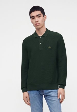 Giovane uomo con capelli corti neri che indossa una polo verde scuro a maniche lunghe e jeans azzurri contro uno sfondo chiaro uniforme.