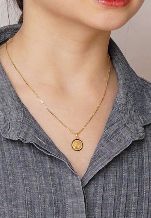 Goldene Anhängerhalskette mit einem kreisförmigen Charme, der ein kompliziertes Design aufweist, aufgehängt an einer zarten goldenen Kette, getragen über einem grauen Hemd mit Kragen.