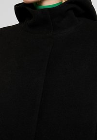 Abrigo de lana negro con un cuello alto, textura suave y diseño minimalista, con un acento verde visible debajo.
