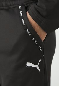 Schwarze Sporthosen mit glatter Textur, ausgestattet mit einem auffälligen weißen Puma-Logo und einem markenbezogenen Bund, der mit dem wiederholten "PUMA"-Text verziert ist.