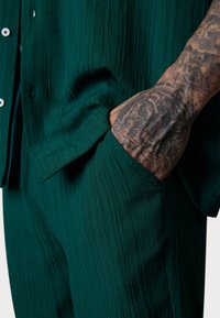 Dark green textured shirt with a pocket, worn by a person with a tattooed hand resting. Focus on the fabric's rippled pattern.