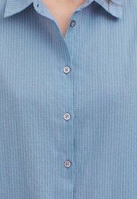 Camicia azzurro chiaro con bottoni, caratterizzata da strisce bianche verticali, un colletto classico e tre bottoni di chiusura sul davanti, con una texture liscia.