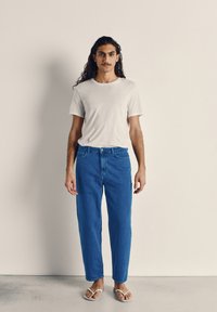 Bela majica s kratkimi rokavi, združena z modrimi kavbojkami z ravnimi nogavicami, ki prikazujejo klasično denim teksturo. Nošeno z belimi japonkami.