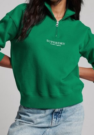Osoba nosząca zieloną bluzę z zamkiem do ćwiartki z napisem "Superdry London, England", połączoną z jasnoniebieskimi jeansami.