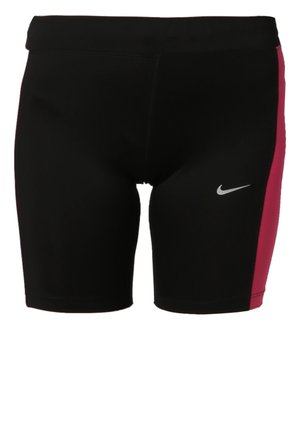 Kobieta idąca w czarnym sportowym topie Nike, czarnych i różowych szortach oraz czarnych tenisówkach na jednolitym jasnym tle.