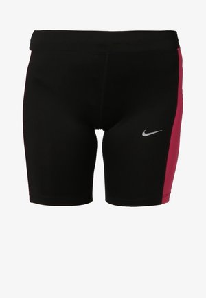 Kobieta idąca w czarnym sportowym topie Nike, czarnych i różowych szortach oraz czarnych tenisówkach na jednolitym jasnym tle.