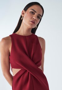 Bezrekawnikowa sukienka w kolorze bordowym o teksturowanym wykończeniu, z zakręconym przodem i wycięciami po bokach. Prosty, okrągły dekolt i eleganckie linie.