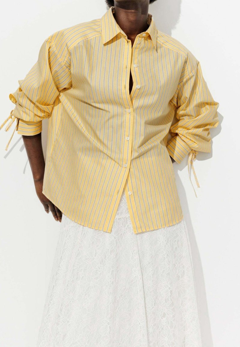 Personne portant une chemise à rayures jaunes boutonnée avec des manches retroussées et une jupe en dentelle blanche sur fond blanc.