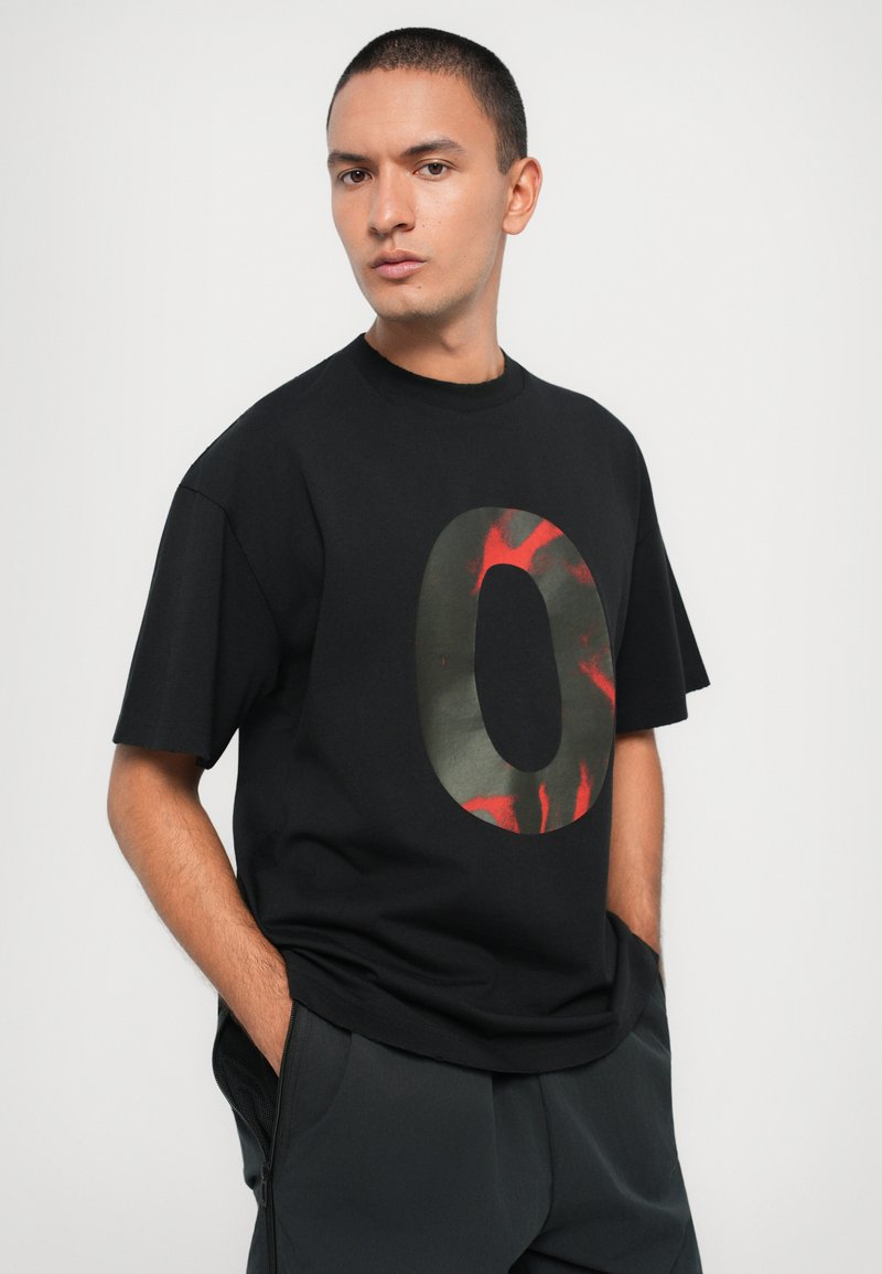 Schwarzes T-Shirt mit kurzen Ärmeln und einem großen, in grün und rot bedruckten "O". Weiches Material, lockere Passform, runder Ausschnitt.