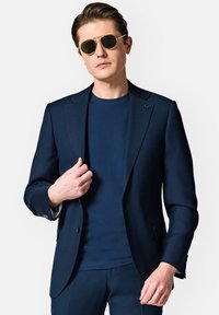Granatowa marynarka o ostrych klapach, noszona na niebieskiej koszuli, w połączeniu z okrągłymi okularami przeciwsłonecznymi; gładka faktura, nowoczesny design, subtelne detale.