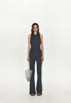Mujer con top sin mangas oscuro de canalé y pantalones acampanados, sosteniendo una bolsa de malla, con gafas de sol y sandalias, sobre un fondo blanco liso.