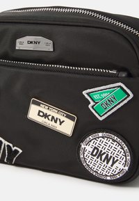 Borsa nera in nylon con zip argentate, tre toppe con marchio e testo ricamato. Forma rettangolare compatta con una tasca frontale.