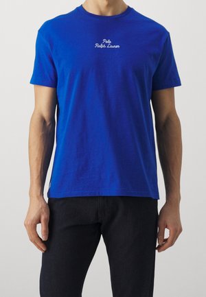 Mężczyzna w jasnoniebieskiej koszulce z krótkim rękawem z białym napisem „Polo Ralph Lauren” na piersi, w połączeniu z czarnymi spodniami.