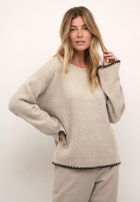 Beżowy, oversize sweter z miękkiego materiału, z czarnymi wykończeniami przy mankietach i dole, o okrągłym dekolcie oraz opadających ramionach.