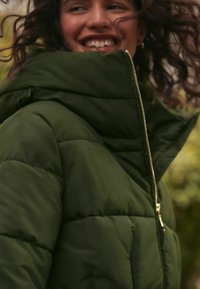 Tmavozelená páperová bunda s vysokým golierom, zipsom a prešívanou textúrou; má priliehavý strih a viditeľné prešívacie detaily.