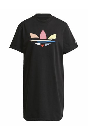 Kvinde iført en sort kortærmet kjole med et flerfarvet Adidas-logo, hvide Adidas-sneakers og sort-hvidt striped ankelsokker.