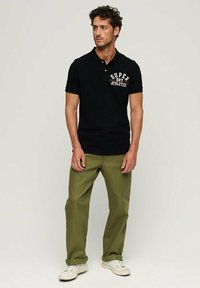 Polo nero con la scritta "SUPER DRY ATHLETICS" stampata in bianco e rosso. Abbinato a pantaloni verde oliva e scarpe da ginnastica bianche. Tessuto dalla consistenza liscia.