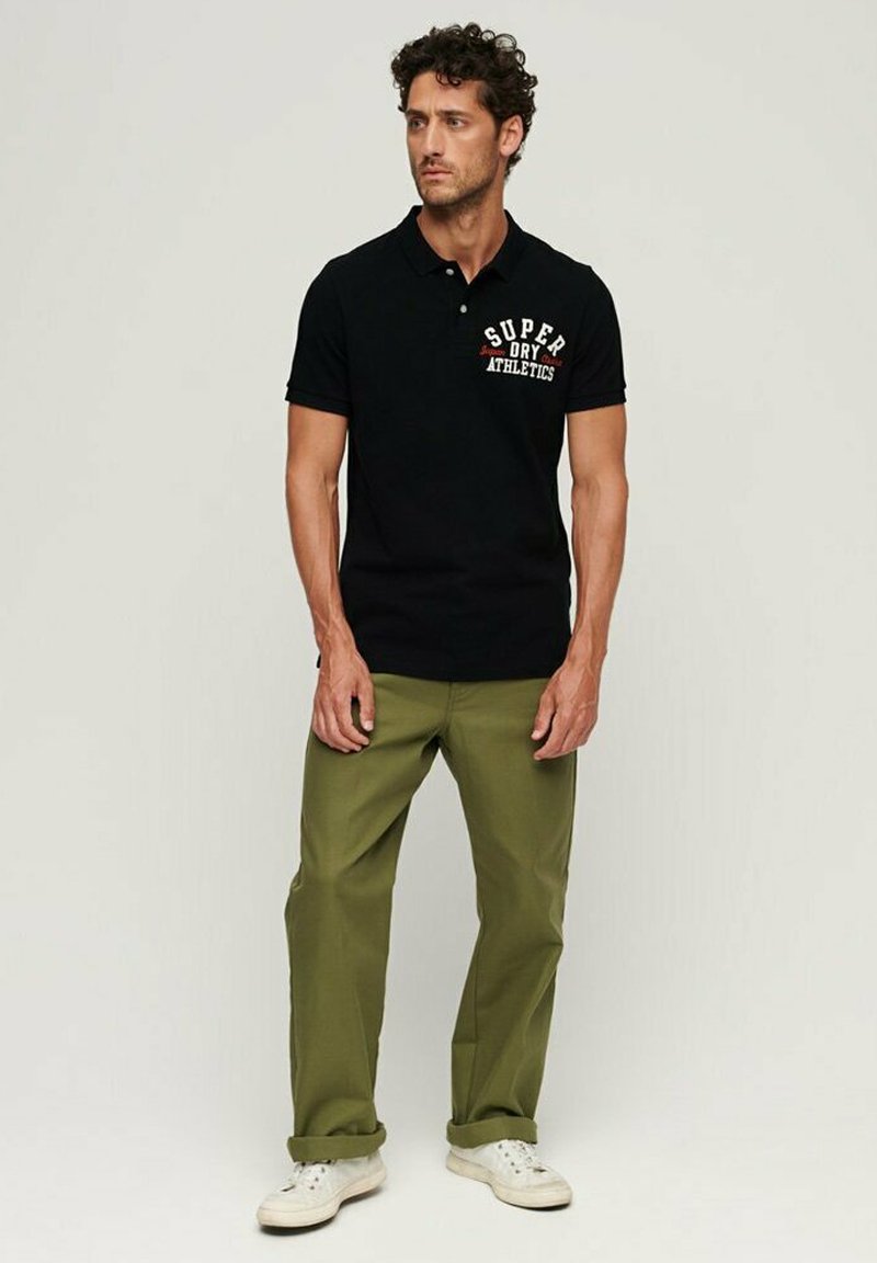 Polo nero con la scritta "SUPER DRY ATHLETICS" stampata in bianco e rosso. Abbinato a pantaloni verde oliva e scarpe da ginnastica bianche. Tessuto dalla consistenza liscia.