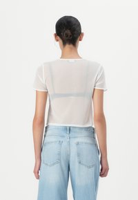 Femme aux cheveux relevés en chignon, portant un haut blanc transparent à manches courtes par-dessus un soutien-gorge noir et un jean taille haute bleu clair, vue de dos.