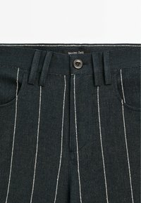 Pantalon en denim foncé avec des rayures blanches verticales, doté d'une fermeture à boutons, de passants pour ceinture et de poches avant. Étiquette de la marque visible sur la ceinture.