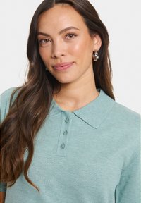 Mentazöld pólóing galléros nyakkal, gombos frontrésszel és finom bordás textúrával. Hosszú haj és ezüst karika fülbevaló látható.