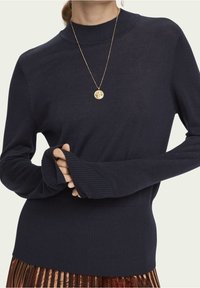 Granatowy sweter dziany z wysokim kołnierzem, prążkowanymi rękawami i dołem; posiada otwory na kciuki. Towarzyszy mu złota kolia z zawieszką.