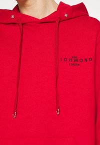 Sudadera roja hecha de una mezcla de algodón que presenta un bolsillo frontal, cordones ajustables con puntas de metal y un logo bordado en el pecho.