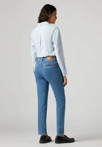 Camicia azzurro chiaro con maniche lunghe abbinata a jeans blu aderenti. I jeans presentano una toppa in pelle marrone e tasche posteriori con dettagli rossi.