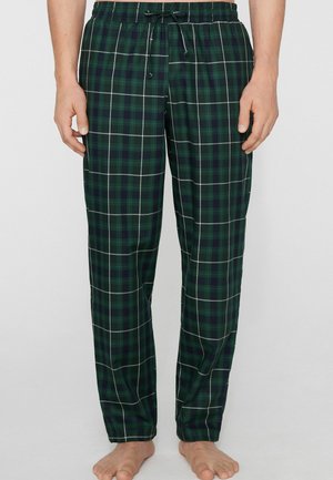 Hombre con pantalones de pijama de cuadros verdes y azul marino con líneas blancas, de pie descalzo frente a un fondo liso y claro.