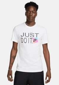 T-shirt in cotone bianco con una grande grafica nera "JUST DO IT." e una patch del logo Nike rosa e blu sulla parte anteriore. Maniche corte.