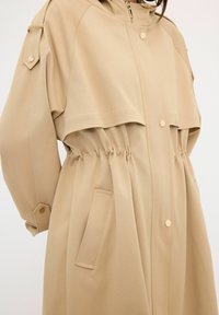 Beige ytterkläder med midja i elastiskt material, med en zip-up-framdel, tryckknappar och sidofickor. Tillverkad av ett smidigt material med huva.