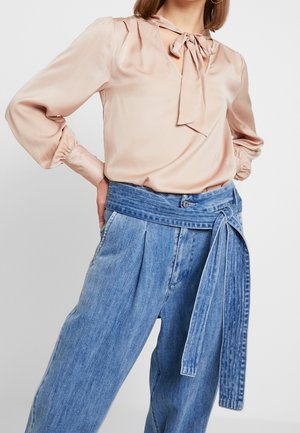 Femme portant un chemisier en satin rose clair avec un col à nouer et un pantalon en jean bleu taille haute avec une large ceinture à nouer.