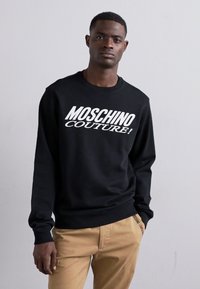 Sweatshirt noir à col ras du cou, présentant un audacieux texte blanc "MOSCHINO COUTURE!", conçu en tissu lisse, avec des poignets et un ourlet côtelés.