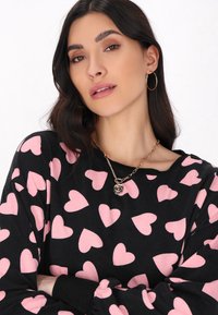 Svart långärmad tröja med rosa hjärtan, med rund halsringning. Guldhalsband med en rund berlock och stora örhängen.