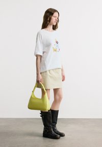Γυναίκα που φοράει λευκό γραφικό μπλουζάκι και μπεζ φούστα κρατάει ένα τσάντα σε λιμονί πράσινο, στέκεται με μαύρες μπότες μέχρι το γόνατο σε γκρι πάτωμα.