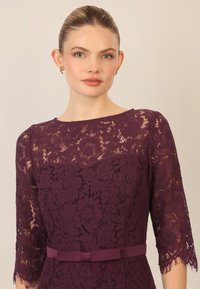 Abito in pizzo prugna con maniche a tre quarti, motivo floreale, scollatura rotonda e una cintura in satin in vita per un tocco di dettaglio.