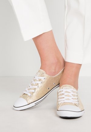 Zapatillas bajas texturizadas en oro con cordones blancos y punteras blancas, llevadas con pantalones blancos recortados sobre un fondo claro.