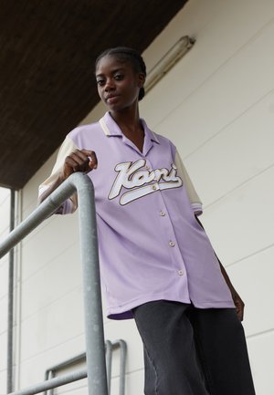 Fialová košile s krátkým rukávem a krémovým límcem, na přední straně s velkým bílým kurzivním logem. Model stojí proti kovovému zábradlí.