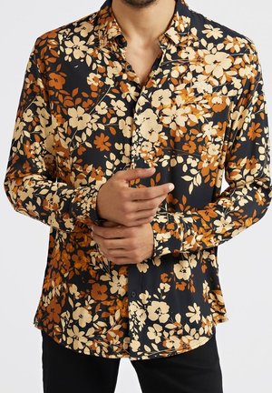 Mężczyzna w czarnej koszuli z długim rękawem, ozdobionej pomarańczowo-kremowym kwiatowym wzorem, zapiętej z przodu na guziki, z rękami złożonymi na wysokości talii.