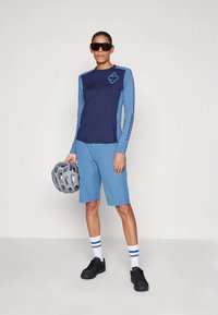 Top atlético de manga comprida em azul-marinho e azul claro, combinado com calções azul claro, óculos de sol e capacete cinzento. Ténis pretos com detalhes brancos.