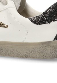 Zapatilla blanca con acentos de purpurina negra y dorada, parte superior de cuero suave, puntera redondeada y suela de goma texturizada con un ligero desgaste.