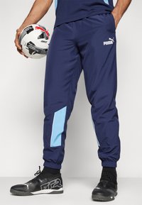Tengerészi sportnadrág világoskék oldalpanellel, rugalmas mandzsettákkal és Puma logóval. Fekete sportcipővel viselve, focilabdát tartva.