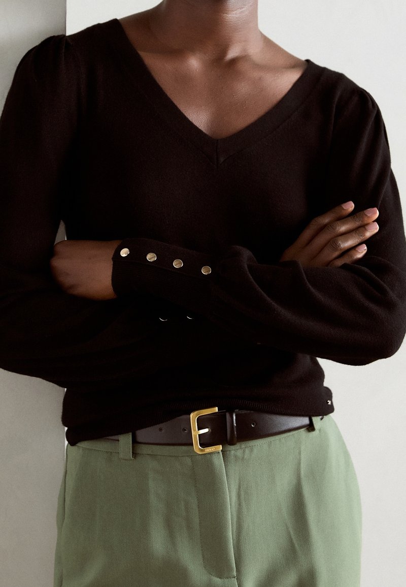 Maglione nero con scollo a V e maniche a sbuffo, caratterizzato da bottoni metallici sui polsini. Abbinato a pantaloni verdi e una cintura nera con fibbia dorata.