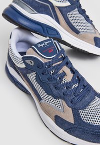 Blå och grå atletiska skor med meshpaneler, mockadetaljer och blå gummisulor. Har runda snören och en logotypetikett på tungan.